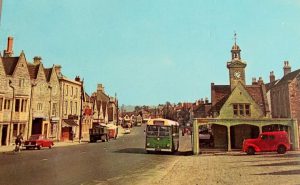 Chipping Sodbury High Street in 1958 with people walking and cycling and vehicles such as a green bus and red cars